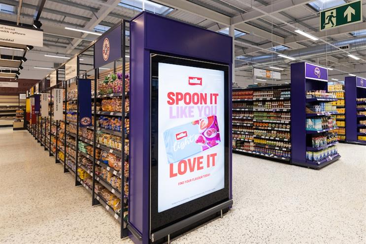 Large purple digital screen at the end of a supermarket aisle running a Müller Light campaign, surrounded by stocked shelves